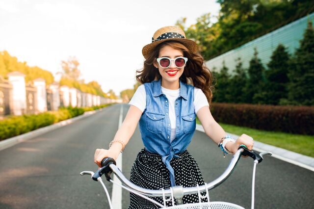 cute-girl-with-long-curly-hair-in-sunglasses-driving-a-bike-to-camera-on-road-she-wears-long-skirt-jerkin-hat-she-looks-happy